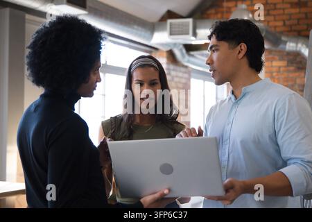 Verschiedene Kollegen, die sich über einen Laptop in einem Open Office beugen, mit Stirnband-Halskette, schicker Kleidung Stockfoto