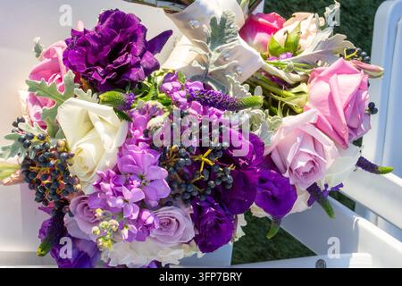 Eine Nahaufnahme eines farbenfrohen Hochzeitsstraußes mit dunkelvioletten Lisianthus, lavendelfarbenen Rosen und Viburnum-Beeren, die auf einem weißen Stuhl ruhen. Stockfoto