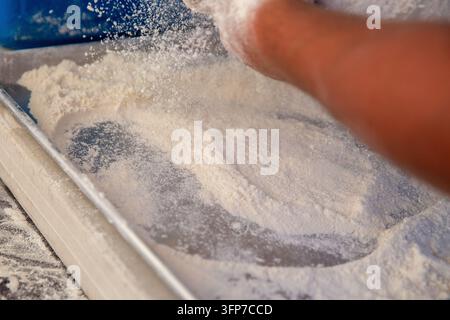 Eine Nahaufnahme einer Action-Szene eines Bäckers, der feines Weißmehl auf eine metallene Backblechpfanne stäubt. Stockfoto