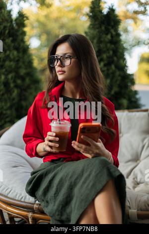 Eine stilvolle junge Frau mit Brille und rotem Hemd sitzt in einem Außenstuhl, hält Eiskaffee und ein Smartphone und schaut nachdenklich in Stockfoto