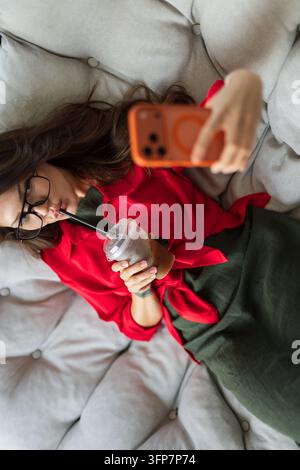 stilvolle Frau in einem roten Hemd, die auf einem gemütlichen Sofa liegt, durch einen Strohhalm Eiskaffee trinkt und mit ihrem Smartphone ein Selfie macht, während sie entspannt. Stockfoto