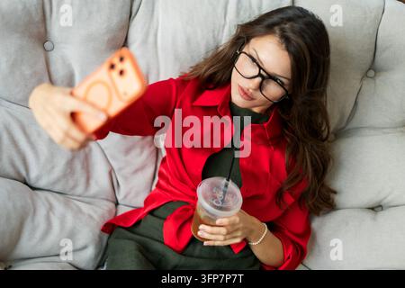 Trendige junge Frau mit Brille und einem hellroten Hemd, die ein Selfie auf ihrem Smartphone macht, während sie einen Eiskaffee hält und auf einem bequemen Sofa liegt Stockfoto