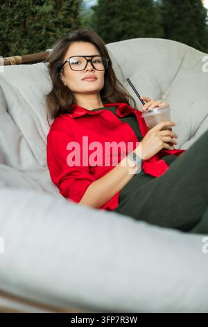 Stilvolle junge Frau in rotem Hemd und Brille, die auf einem gemütlichen Außenstuhl entspannt, direkt in die Kamera schaut und einen Eiskaffee hält. Stockfoto