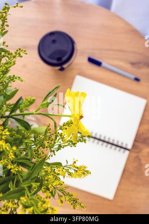 Eine Nahaufnahme von gelben Blumen in einer Vase im Fokus. Ein unscharfer Hintergrund mit einem Notizblock, einem Stift und Kaffee. Eine gemütliche Heimbüro-Szene mit natürlichem Licht. Stockfoto
