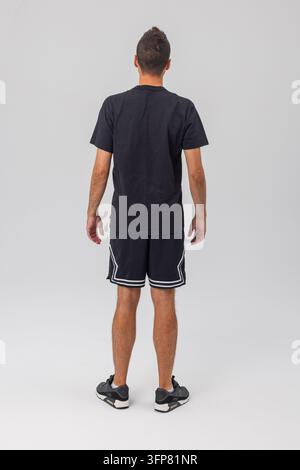 Studiofoto: Rückansicht eines muskulösen männlichen Models, das ein schlichtes schwarzes Baumwoll-T-Shirt und athletische Shorts mit weißen Streifen trägt. Isoliert auf Grau. Stockfoto