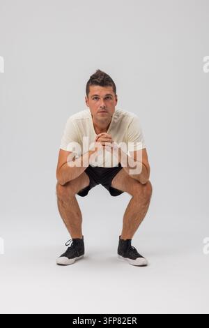 Ein junger Mann hockt auf einem neutralen grauen Hintergrund tief, schaut in die Kamera mit verschränkten Händen. Er trägt ein weißes T-Shirt und schwarze Shorts. Sauberes Studio-Schuh Stockfoto