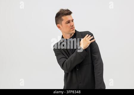Stylischer Mann in einer schwarzen Hemdjacke, der auf seine Brust hinunterschaut und Staub abklopft, isoliert vor hellgrauem Studiohintergrund. Modernes Männerporträt mit Stockfoto
