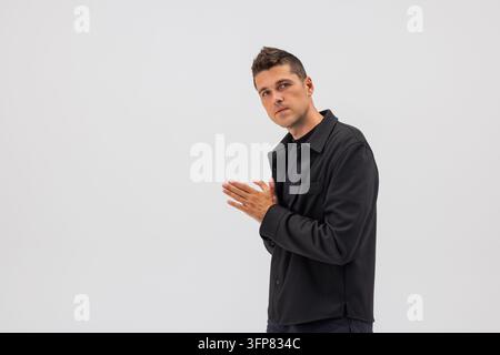 Selbstbewusster junger Mann in schwarzer Jacke mit verschränkten Händen, blickt in die Kamera. Modernes Männerporträt, isoliert auf hellgrauem Hintergrund, ideal für Geschäfte Stockfoto