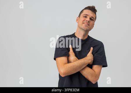 Selbstbewusster junger kaukasischer Mann in einem lässigen schwarzen T-Shirt, posiert mit verschränkten Armen vor einem schlichten hellen Studiohintergrund. Freundliches männliches Porträt mit c Stockfoto
