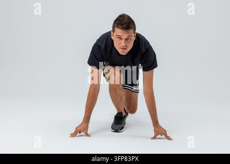 Athletischer Mann in Sportkleidung, der von einer geduckten Position aus einen Sprint beginnt vor einem hellen Studiohintergrund, der Geschwindigkeit, Kraft, Fitness, Training demonstriert, Stockfoto