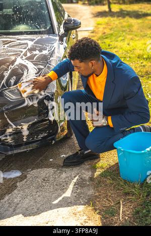 Gut gekleideter junger Mann kniet neben einem schwarzen Auto und schrubbt die schaumige Frontplatte mit einem Schwamm auf einer sonnigen Einfahrt, mit blauem daneben, was eine klare Szene des h erzeugt Stockfoto