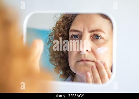 Mittlere Frau, die im Spiegel Gesichtscreme aufträgt und sich auf Hautpflege und Alterungsvorbeugung konzentriert Stockfoto