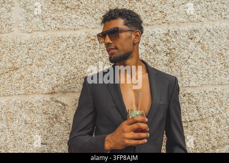 Porträt eines jungen Mannes mit gestutztem Bart, der einen offenen schwarzen Blazer, getönte Sonnenbrille und einen kleinen Ohrring trägt und ein rotes Getränk an einer rauen Steinmauer hält Stockfoto