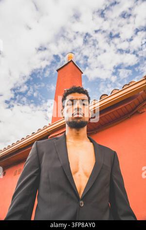 Selbstbewusster junger Mann mit kurzen lockigen Haaren und Bart posiert im Freien neben lebhafter orangefarbener Architektur, von unten betrachtet vor einem hellen bewölkten Himmel, für Stockfoto
