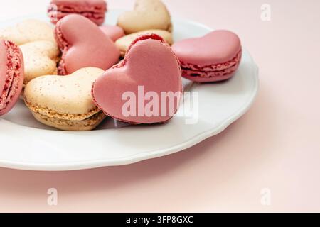 Nahaufnahme von rosa und Vanille-Herz-Makronen auf einem weißen, muschelförmigen Teller, Fokus auf einem rosa Herz, hellrosa Hintergrund. Stockfoto