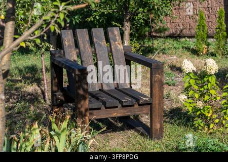 Handgefertigter dunkler Holzstuhl in einem sonnigen grünen Garten, Nahaufnahme. Rustikaler Holz-Adirondack-Stuhl auf dem Rasen im Hinterhof. Bequemes Outdoor-Möbelset in Sum Stockfoto