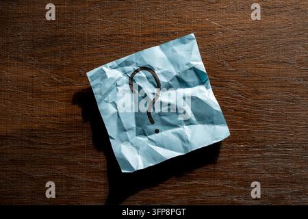Fragezeichen auf zerknittertem blauem Papier notiert auf Holzuntergrund. Zerknitterter blauer Klebezettel mit handgeschriebenem schwarzem Fragezeichen. Konzept von Verwirrung Stockfoto