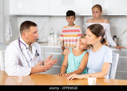 Während einer Beratung vor Ort zu Hause erzählt ein männlicher Arzt der Mutter mit vielen Kindern von der Behandlung eines weiblichen Kindes. Die kranke Tochter sitzt auf der Mutter Stockfoto