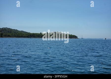 Kroatien Ruhiges blaues Meer mit bewaldeter Inselküste unter klarem Himmel, mit entferntem Segelboot und kleinem Schnellboot auf offenem Wasser. Weiter Horizont und sonnenbeschienene r Stockfoto
