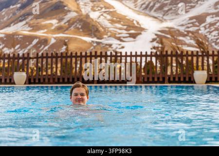 Mittelformat-Aufnahme eines lächelnden gutaussehenden Mannes oder Teenagers, der in einem luxuriösen Winterberg-Spa-Resort-Pool entspannt, mit schneebedeckten Pisten im Hintergrund. Stockfoto