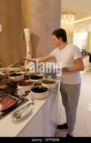 Vertikales Porträt eines gutaussehenden Mannes, der Oliven und Snacks an einem Premium-Resort-Restaurant-Buffet am Morgen nimmt. Stockfoto