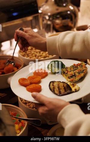 Nahaufnahme einer vertikalen Aufnahme von Händen, die einen weißen Teller mit Brokkoli, gegrillter Aubergine und Omelett in einem Premium-Resort-Restaurant-Buffet halten. Stockfoto
