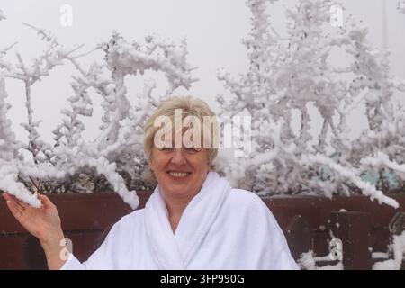 Lächelnde reife Frau in einem weißen Bademantel entspannt sich in einem luxuriösen Winter-Wellness-Spa-Resort mit schneebedeckten Bäumen im Hintergrund. Stockfoto