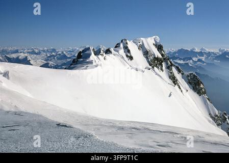 Ein majestätischer schneebedeckter Berggipfel dominiert die Szene unter einem klaren blauen Himmel. Die weitläufige Landschaft zeigt sanfte Hügel und entfernte Gipfel fadi Stockfoto