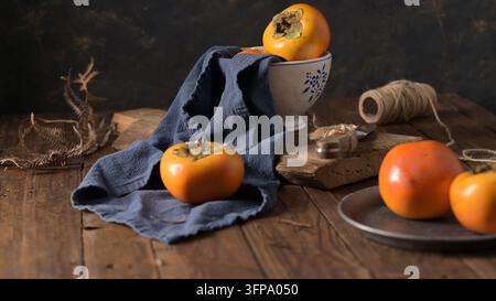 Eine Nahaufnahme von lebhaft orangefarbenen Persimmons, die mit einem drapierten blauen Tuch, einer Keramikschale und einem kleinen Metalltablett auf einer dunklen Holzoberfläche stilisiert sind, beleuchtet Stockfoto