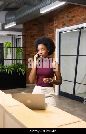 Afrikanische Frau in einem burgunderroten Oberteil, die hinter dem Empfangstresen im Büro steht und ein Telefon neben einem Laptop hält Stockfoto