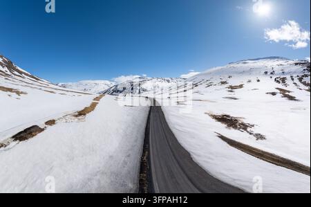 Blick auf eine leere Asphaltstraße, die sich durch eine weite, schneebedeckte Landschaft unter einem strahlend blauen Himmel in Island schlängelt Stockfoto