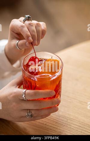 Nahaufnahme der Hände einer Frau, die eine Cocktailkirsche in einem Glas rotem Negroni als Garnitur mit Orangenschale anpasst. Stockfoto