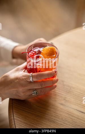 Frau hält ein Glas mit einem roten Cocktail, garniert mit einer Orangenschalenspirale und einer Cocktailkirsche über einem Holztisch. Stockfoto