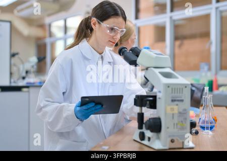 Junge erwachsene Wissenschaftlerin in einem weißen Laborkittel und einer Schutzbrille, die auf ein digitales Tablet schaut, während sie biologische Forschung mit einem Mikroskop durchführt Stockfoto