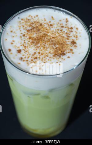Ein hohes Glas erfrischender Eistee-Latte aus Matcha-Grüntee, gekrönt mit dickem weißem Milchschaum und bestreut mit aromatischem Gewürzpulver vor schwarzem Hintergrund Stockfoto
