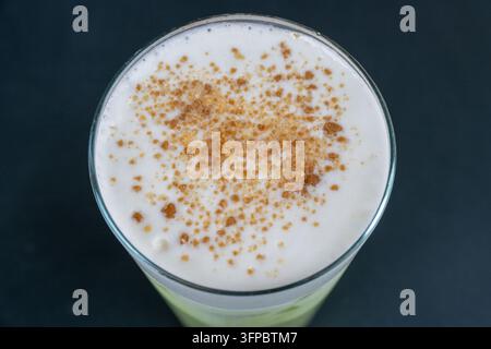 Ein Glas Getränk, das mit dickem weißem Milchschaum gekrönt und mit braunem gewürztem Zuckerpulver bestreut ist, vor einem dunklen Hintergrund. Stockfoto