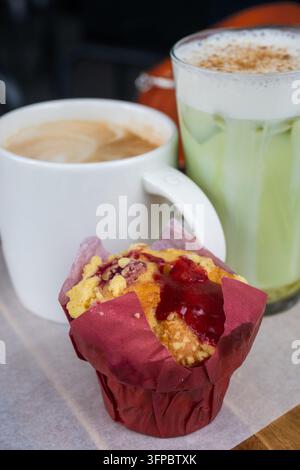 Ein köstliches Café-Frühstücks-Arrangement mit einem frisch gebackenen Beeren-Crumble-Muffin in einer roten Tulpenverpackung, gepaart mit einem eisgekühlten Matcha-Latte mit einem Topping aus Stockfoto