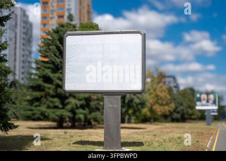 Ein leerer Outdoor-Werbeplakat-Mockup auf einem Metallpfosten, der auf einer urbanen Parkwiese mit Bäumen und Stadtgebäuden im Hintergrund steht. Stockfoto