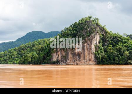Ein schlammiger brauner Fluss fließt an einem großen Kalkstein-Karst vorbei, der in üppiger grüner Vegetation nahe Luang Prabang, Laos, bedeckt ist. Stockfoto