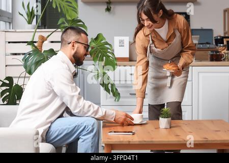 Kundenservice und Gastfreundschaft, Barista serviert Kaffee und Gebäck an einen männlichen Kunden in einem modernen Café mit Pflanzen und gemütlichem Innendesign Stockfoto