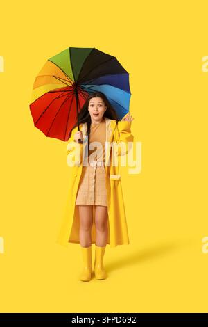 Wettervorhersage und Vorbereitung auf die Regensaison, überraschte junge Frau hält einen bunten Regenbogen-Regenschirm und trägt einen gelben Regenmantel vor einem gelben Hintergrund Stockfoto