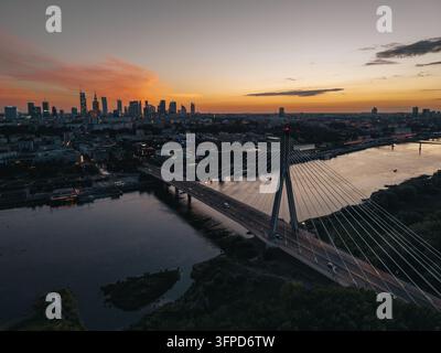 Warsaw Skyline bei Sonnenuntergang mit der Swietokrzyski-Brücke, die die Weichsel überspannt, lebendiges Abendlicht und streifende Autoscheinwerfer, die sich auf dem Wasser spiegeln Stockfoto