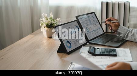 Geschäftsmann analysiert Finanzdaten mit Tabellenkalkulationen auf Laptop und Tablet, zeigt mit einem Stift neben einem Taschenrechner und gedruckten Berichten für die Finanzüberprüfung Stockfoto