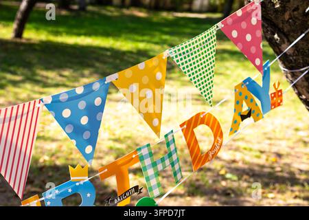 Bunte dreieckige Wimpelflaggen und die letzten Buchstaben eines Geburtstags hängen zwischen Bäumen im klaren Sonnenlicht. Punkte und Streifen heben sich von einem grünen Hintergrund ab Stockfoto