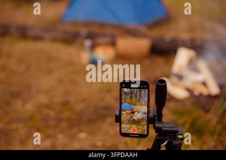 Smartphone auf Stativ montiert, das Video von Lagerfeuer und Zeltaufbau auf einem Herbstwald-Campingplatz aufnimmt Stockfoto
