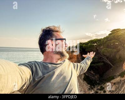 Mann macht ein freudiges Selfie auf einer Küstenklippe bei Sonnenuntergang mit Blick auf das Meer, Arme ausgestreckt, genießt die Natur, das Reisen und die Freiheit mit einem Bart und Su Stockfoto