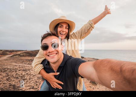 Junges Paar lächelt und lacht, während Männer Frauen auf dem Rücken tragen und die Freiheit und das Glück während eines fröhlichen Sommerurlaubs am Meer feiern Stockfoto