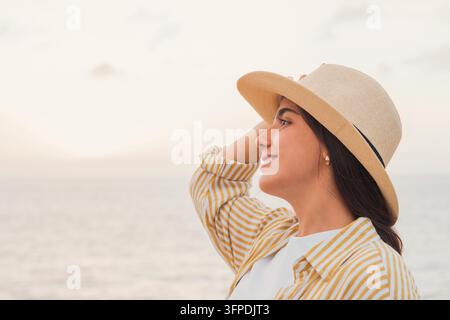 Junge Frau lächelt, während sie in die Ferne blickt, schützt die Augen mit einer Hand, trägt einen Hut, fühlt sich unbeschwert und genießt einen friedlichen Moment am offenen Meer Stockfoto