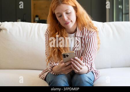 Junge rothaarige Frau sitzt auf einem weißen Sofa zu Hause, lässig gekleidet in einem gestreiften Hemd und Jeans, schaut nachdenklich auf ihr Smartphone und vermittelt Entspannung, Konzentration und ruhiges Alltagsleben. Stockfoto