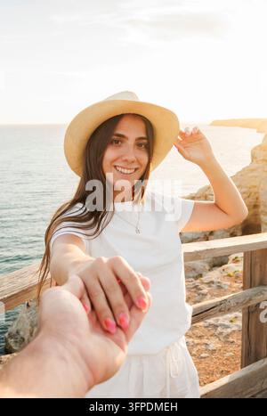 Frau mit Strohhut und weißem Outfit lächelt und greift nach hinten, um Sie entlang einer sonnenbeschienenen Küstenklippe zur goldenen Stunde zu führen, und lädt zu Reisen, Romantik und Meeresabenteuern ein Stockfoto
